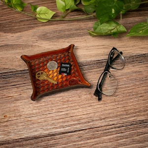 May include: A square brown leather valet tray with a woven texture pattern. Inside the tray are a silver-coloured coin, a brass-coloured key, and a black SanDisk memory card. Next to the tray are black-framed spectacles.