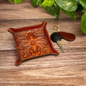 May include: A square brown leather valet tray featuring a deeply embossed floral pattern in the centre and raised corners. Next to the tray lies a metal key attached to a brown leather keychain fob with a gold-toned metal ring.