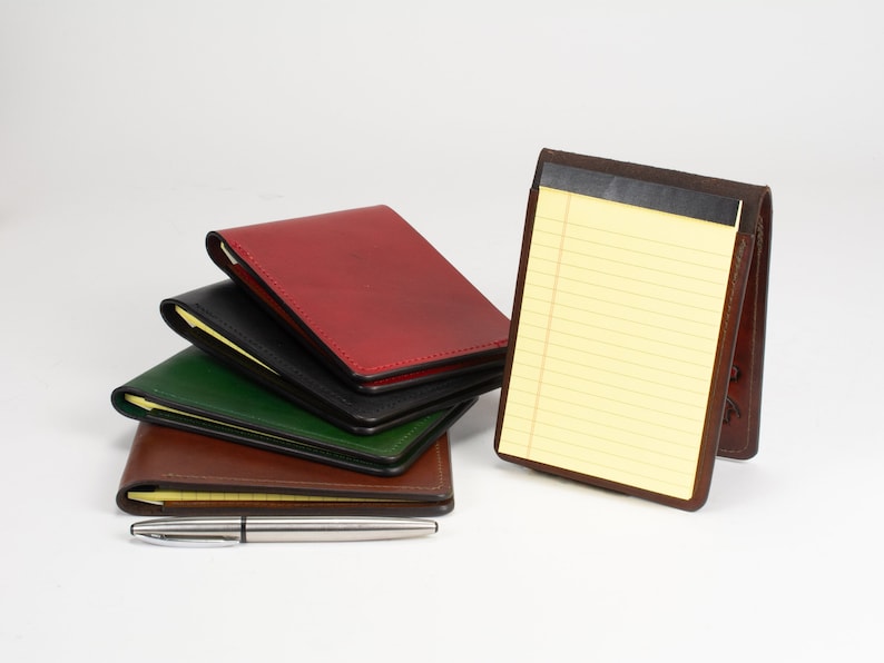 May include: A stack of four leather notepad holders in red, black, green, and brown, with one brown leather holder standing upright containing a yellow lined notepad, accompanied by a silver metal pen placed horizontally in the foreground.