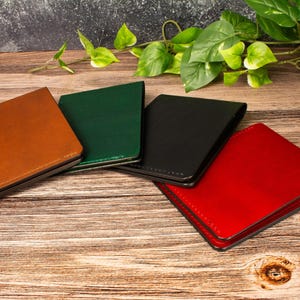 May include: Four rectangular leather passport holders or wallets arranged in a fan shape. The items are colored in tan, dark green, black, and bright red. Each piece features visible stitching along the edges and a smooth, uniform surface finish.
