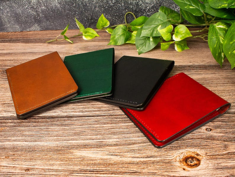May include: Four rectangular leather passport holders or wallets arranged in a fan shape. The items are colored in tan, dark green, black, and bright red. Each piece features visible stitching along the edges and a smooth, uniform surface finish.