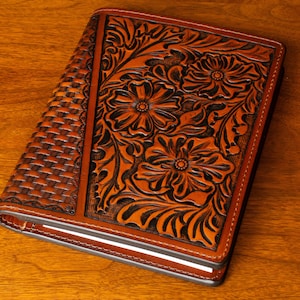 May include: A brown leather journal with a tooled floral design. The cover has a woven pattern on the left side and a floral design on the right side.