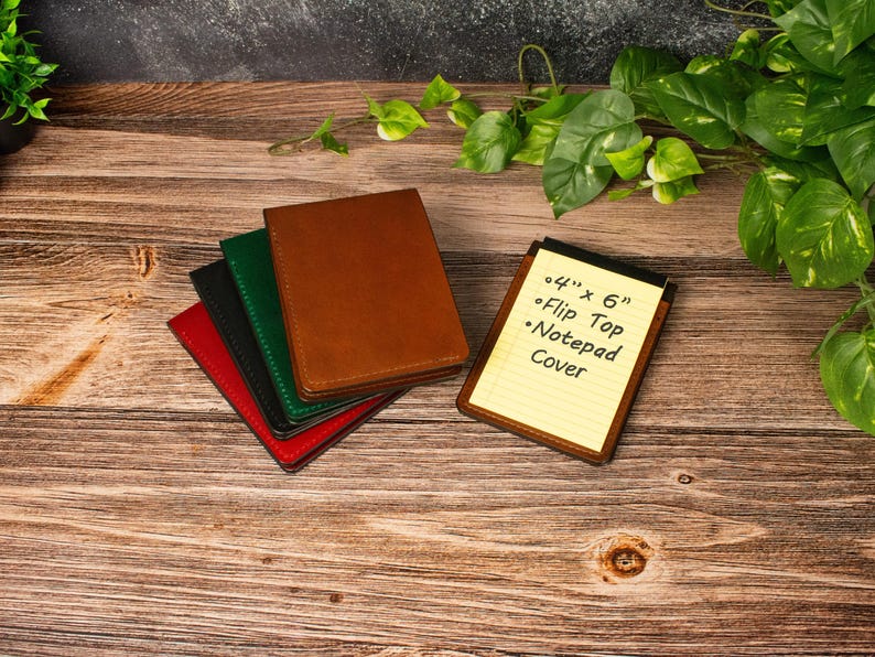 May include: A stack of four rectangular notepad covers in brown, green, black, and red, alongside a single brown flip top notepad cover containing a yellow lined notepad with the text 4" x 6" Flip Top Notepad Cover written on it.
