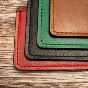 May include: A stack of four rectangular leather card holders in red, black, green, and brown, each featuring rounded corners and visible perimeter stitching in a matching thread color, layered neatly to display the edges of each individual piece.
