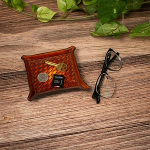 May include: A square brown leather valet tray with a woven texture pattern containing a gold-coloured key, a silver coin, and a black SanDisk memory card, positioned next to a pair of black-rimmed spectacles.