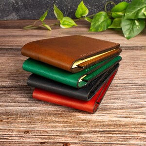 May include: A stack of four bifold wallets in brown, green, black, and red. Each wallet features a smooth, matte finish with visible stitching along the edges and a slim profile. The wallets are layered on top of each other in a vertical arrangement.