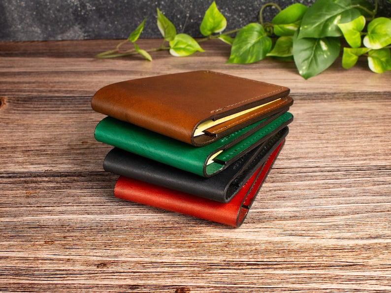 May include: A stack of four bifold wallets in brown, green, black, and red. Each wallet features a smooth, matte finish with visible stitching along the edges and a slim profile. The wallets are layered on top of each other in a vertical arrangement.