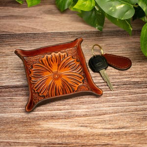 May include: A square brown leather valet tray featuring a deeply embossed floral pattern in the centre. The tray has raised, curved corners and a smooth, polished finish. A metal key on a ring with a brown leather tag rests beside it.