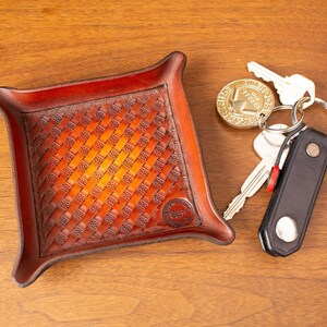 May include: A square, reddish-brown leather tray with a woven pattern, holding keys and a keychain. The tray has a raised border and rounded corners. A brass coin with text is also present.