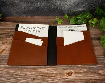 May include: A brown leather four-pocket folder with a black spine, featuring two interior pockets on each side. The folder contains white paper inserts with the printed text Four Pocket Folder and For US Letter Paper, along with small white cards.