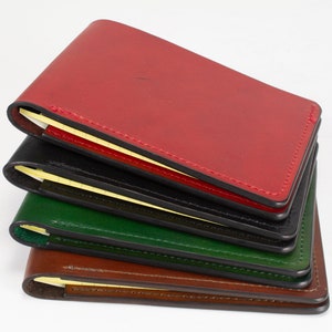 May include: A stack of four rectangular leather notebook covers in red, black, green, and brown. Each cover features visible stitching along the edges and holds a small yellow notepad inside, with the pages visible along the spine of each item.