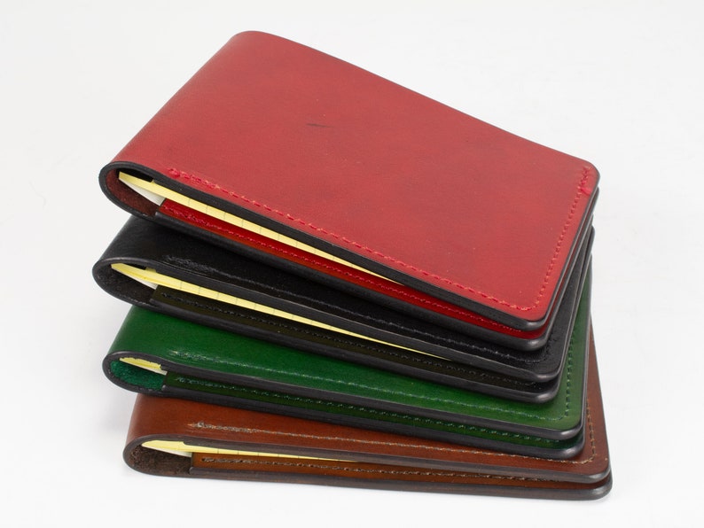 May include: A stack of four rectangular leather notebook covers in red, black, green, and brown. Each cover features visible stitching along the edges and holds a small yellow notepad inside, with the pages visible along the spine of each item.