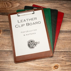 Puede incluir: Un conjunto de cuatro portapapeles de cuero en marrón, verde, negro y rojo. El portapapeles marrón superior sostiene una hoja de papel blanca con el texto LEATHER CLIP BOARD FOR US LETTER & A4 PAPER y un logotipo circular con diseño de hoja de roble.