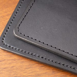 May include: A black leather item featuring smooth, matte surfaces with rounded corners and thick, dark stitching along the edges. The object consists of two layered rectangular pieces, each finished with precise, uniform thread work.
