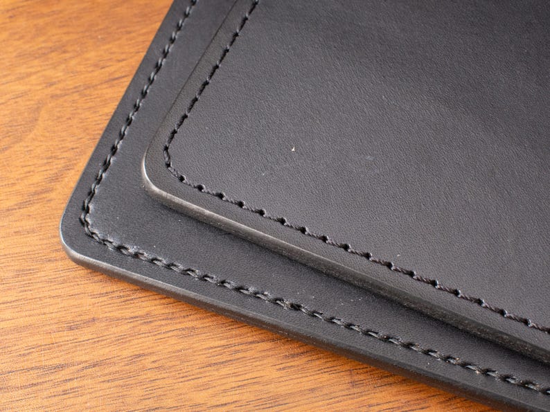 May include: A black leather item featuring smooth, matte surfaces with rounded corners and thick, dark stitching along the edges. The object consists of two layered rectangular pieces, each finished with precise, uniform thread work.