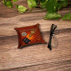 May include: A square brown leather valet tray with a woven texture pattern. Inside the tray are a silver coin, a gold-coloured metal key, and a black SanDisk memory card. Beside the tray sits a pair of black-framed spectacles.