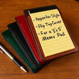May include: A collection of leather memo pad covers in various colors: brown, red, black, and green. A silver pen rests beside the covers. The top cover holds a yellow notepad with handwritten text: "Reporter Style, Flip Top Cover, For a 3"x5" Memo Pad."