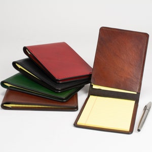 May include: A collection of leather notepad holders in red, black, green, and brown, with one brown holder standing open to reveal a yellow lined notepad. A silver metal pen is positioned to the right of the open notepad holder.
