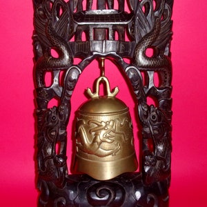 May include: A brass bell with dragon motifs hangs within a dark wooden frame. The frame features carved dragons and a pagoda-style top. The bell is suspended from a gold-colored hanger, creating a striking contrast against the dark wood.