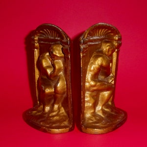 May include: A pair of gold-colored bookends featuring a sculpted design of a seated figure. The bookends are made of metal and have a detailed, ornate design.