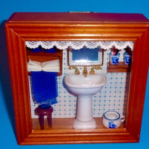May include: Miniature bathroom diorama in a wooden frame. The scene includes a white pedestal sink with gold fixtures, a mirror, and blue and white patterned wallpaper. A blue towel hangs on a small shelf, and a small stool is in front of it.