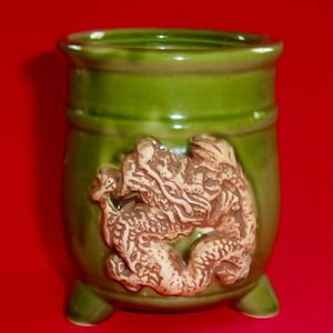 May include: A cylindrical green ceramic vessel featuring a raised, light-colored relief of a dragon wrapped around the exterior. The container has a smooth, glossy finish and is supported by three small, integrated feet at the base.