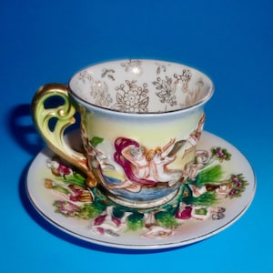 May include: A vintage teacup and saucer set. The cup features a gold handle and is decorated with a classical scene in shades of red, yellow, and white. The saucer mirrors the cup's design, with a gold rim and floral accents.