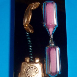 May include: A decorative black plaque featuring a gold-colored telephone and an hourglass with pink sand. The telephone receiver is connected to the base with a green coiled wire. The hourglass is made of clear glass.