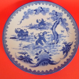 May include: A blue and white porcelain plate with a decorative design of children playing in a garden with a tree and butterflies. The plate has a blue and white border.