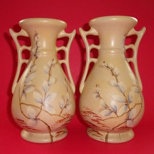 May include: Pair of vintage ceramic vases with a cream background and painted floral design. The vases have a unique shape with two handles on each side.