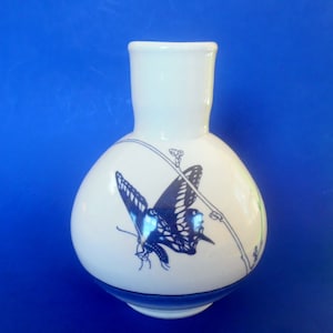May include: A white ceramic vase with a blue and black butterfly design. The vase has a round body and a narrow neck.