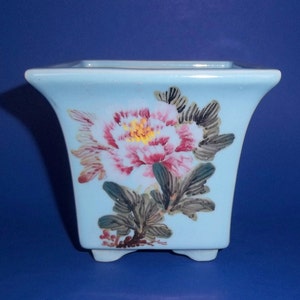 May include: A light blue square ceramic planter with a painted floral design. The flower is pink with a yellow center and green leaves.