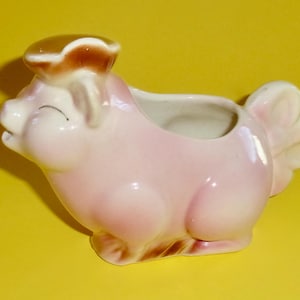 May include: A pink ceramic piggy bank with a brown hat and a small opening on the back for coins. The piggy bank is shaped like a pig with a curly tail.