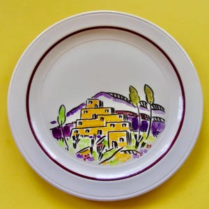 May include: White ceramic plate with a maroon rim and a colorful illustration of a Southwestern village. The design features yellow buildings, purple mountains, and green trees. A decorative piece for the kitchen or dining room.