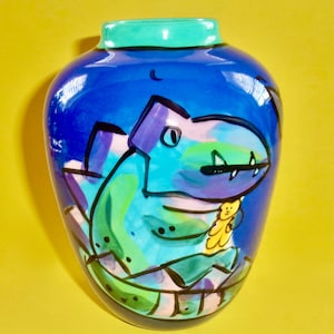May include: A blue ceramic vase with a hand-painted design of a green and purple dinosaur holding a yellow teddy bear.