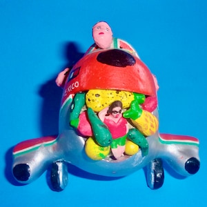 May include: A silver airplane figurine with a red, white, and green design. The airplane is filled with colorful fruit and vegetable figurines, including a pineapple, a green pepper, and a red tomato. The airplane has a small figure of a person with a red shirt and black hair.