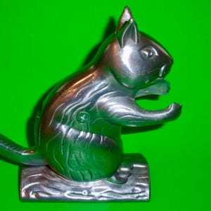 May include: A silver-colored squirrel nutcracker with a detailed design. The squirrel is posed on a log base, with its mouth open, ready to crack nuts. The nutcracker is made of metal and has a shiny finish.