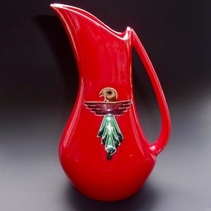 May include: A vibrant red ceramic pitcher with a curved handle and spout. The pitcher features a decorative emblem of a bird with outstretched wings in brown, maroon, and green. The pitcher is a decorative item.