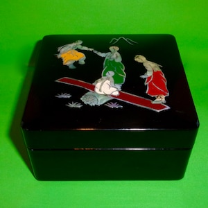 May include: Black lacquered wooden box with a colorful scene of people on a seesaw. The box is decorated with inlaid mother-of-pearl and features a mountain scene in the background.