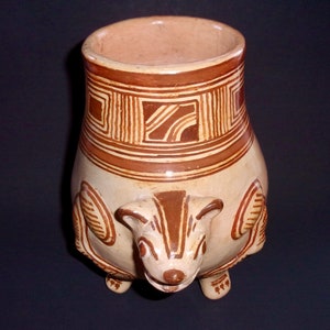 May include: A ceramic vase with a brown and white geometric design and a stylized animal figure on the bottom. The vase has a wide mouth and a rounded body.