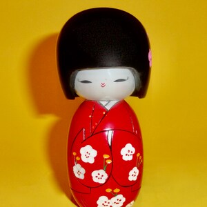 May include: A traditional Japanese kokeshi doll with black hair, a red kimono with white flower designs, and pink flower accents on the hair.