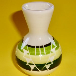 May include: A small white ceramic vase with a green and white geometric pattern. The vase has a narrow neck and a round body.