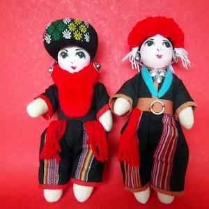 May include: Two handmade dolls wearing traditional clothing. The doll on the left is wearing a black and red outfit with a green and yellow patterned hat. The doll on the right is wearing a black and red outfit with a red hat.