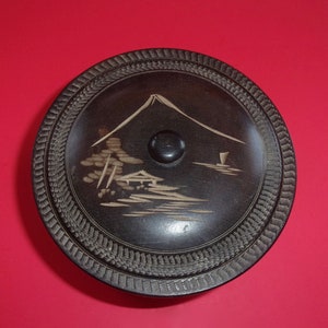 May include: A round, black wooden box with a carved design of a mountain and trees. The box has a black knob on the top.