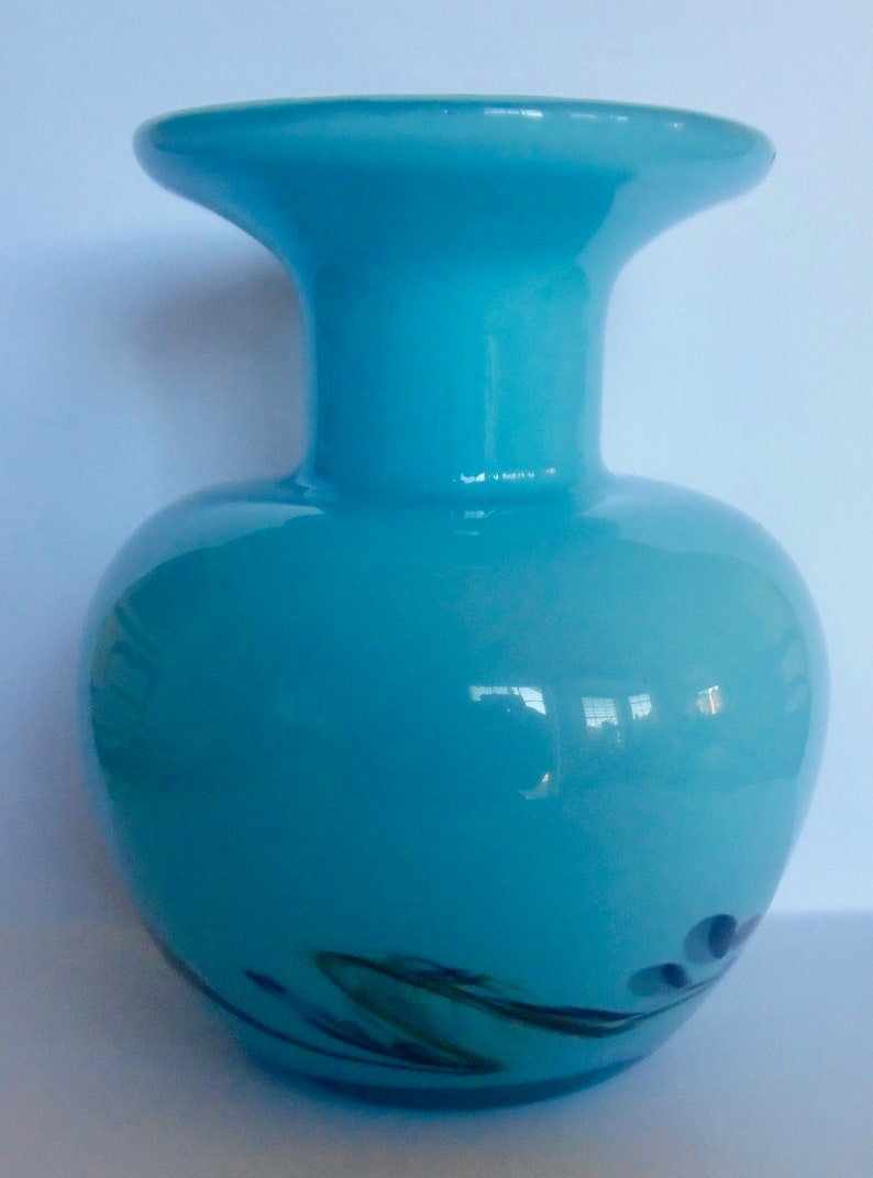 Peut inclure: Un vase en verre bleu clair avec une large ouverture et un corps arrondi. Le vase a un motif en spirale dans des tons de vert et de noir.