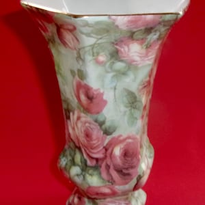May include: A decorative porcelain vase with a flared top and a hexagonal base. The vase is painted with a floral pattern of pink roses and green leaves on a light green background. The rim is trimmed with gold.