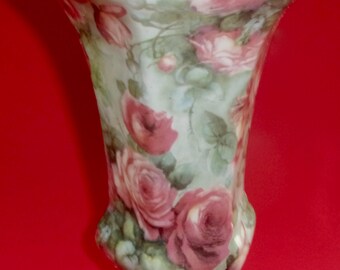 May include: A decorative porcelain vase with a flared top and a hexagonal base. The vase is painted with a floral pattern of pink roses and green leaves on a light green background. The rim is trimmed with gold.