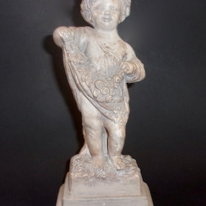 May include: A white ceramic statue of a cherub holding a basket of flowers. The statue is standing on a white pedestal.