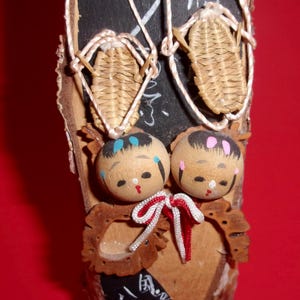 May include: A wooden plaque with two small dolls and two woven straw shoes hanging from it. The dolls have painted faces and the plaque has Japanese writing on it.
