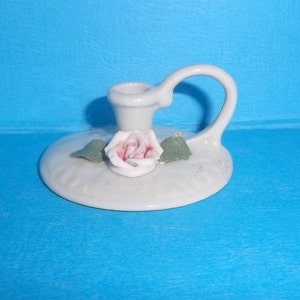 May include: A white porcelain candle holder with a single pink rose and green leaves. The holder has a handle and a round base.